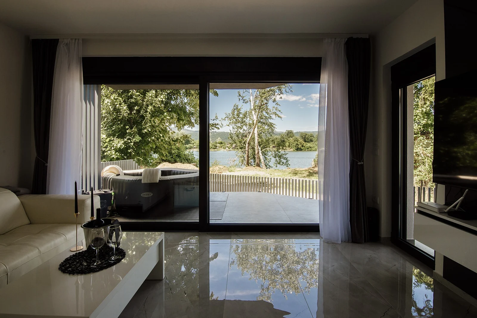 A bright living space with full-height glazing opening toward a wooded lakeside.